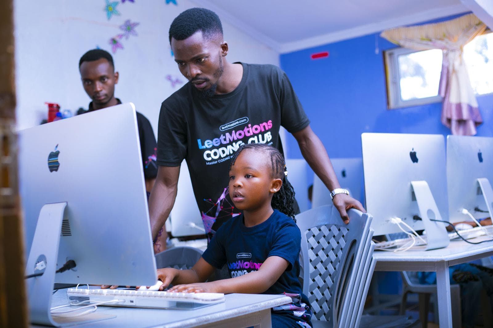 An instructor guiding a young learner, with a row of iMacs behind them